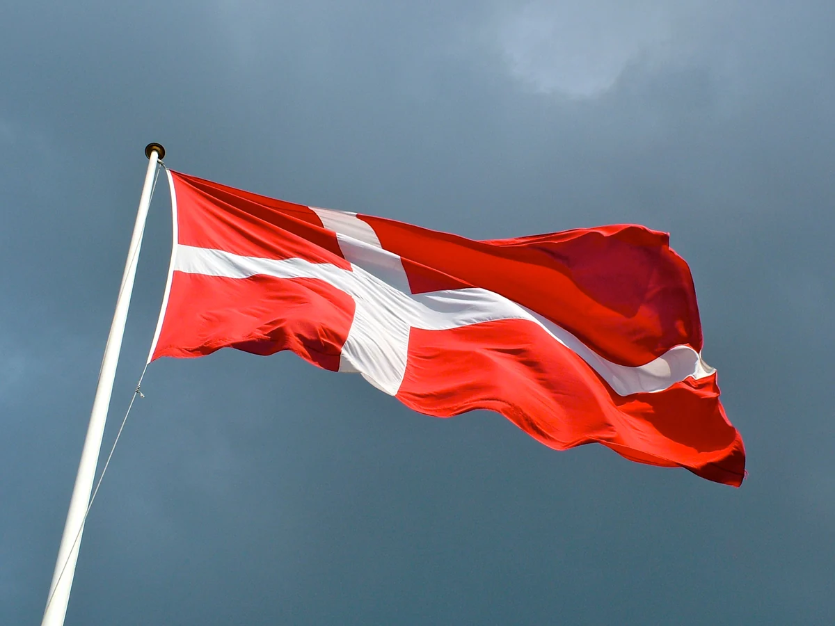 A rectangular Dannebrog flying against a blue sky; the folds show the white cross and red cloth.