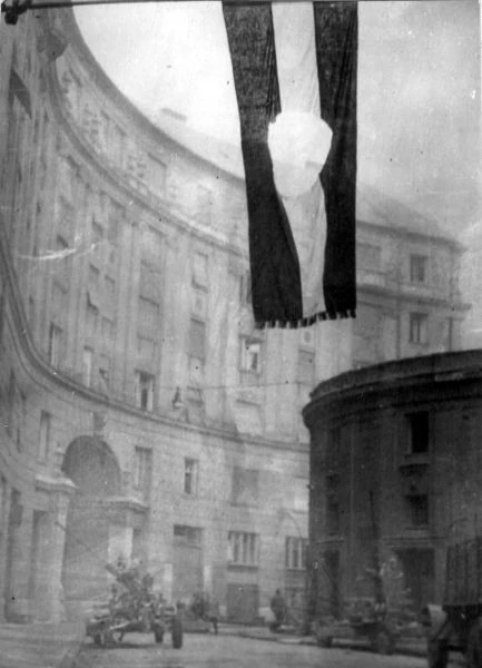 A Hungarian flag with its central emblem cut out hangs above Corvin köz in Budapest during the October 1956 uprising. This archival black-and-white photograph records an actual irregular opening; it cannot establish the original cloth’s exact colors.