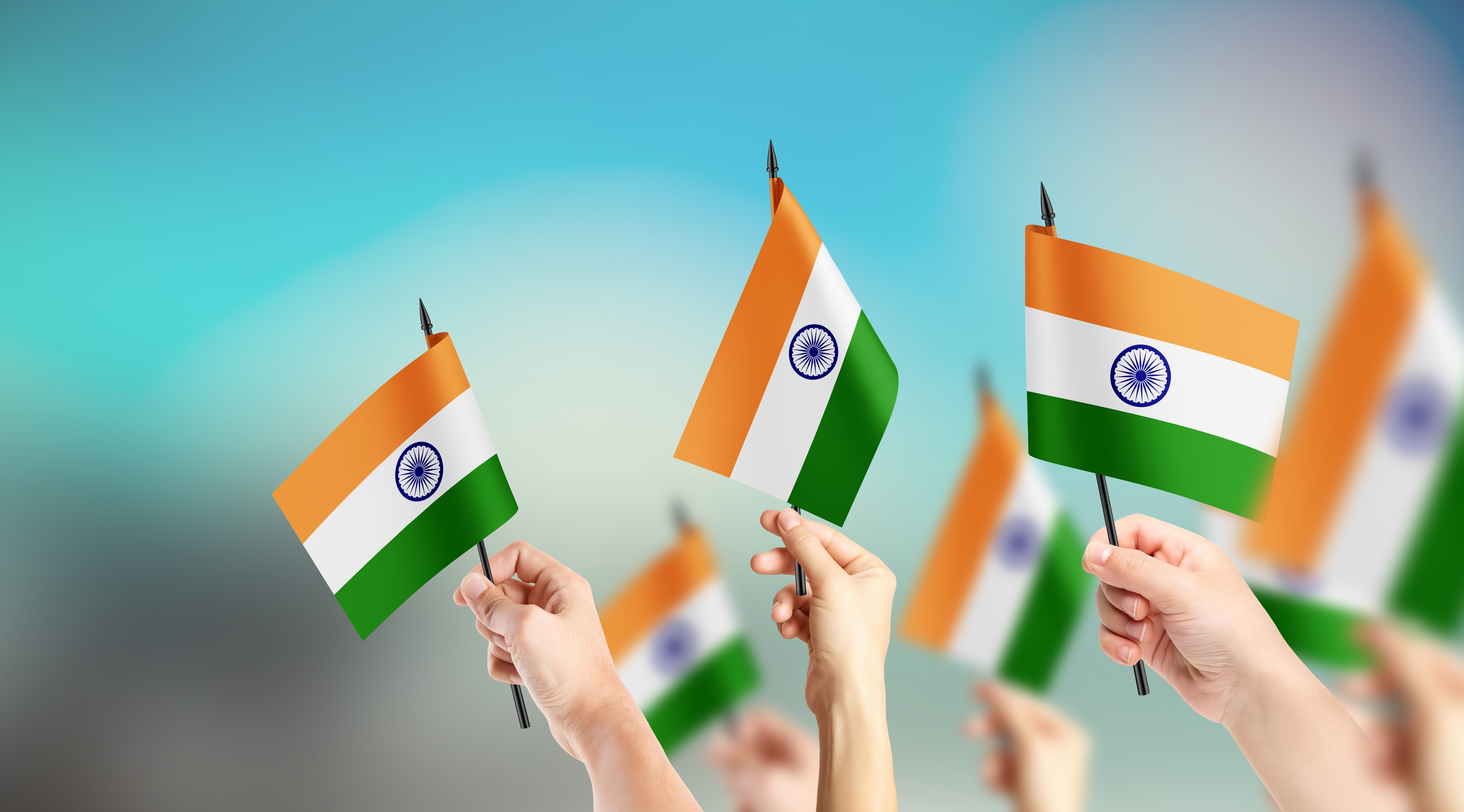 Many raised hands waving small hand-held Indian flags against a blurred blue-green sky